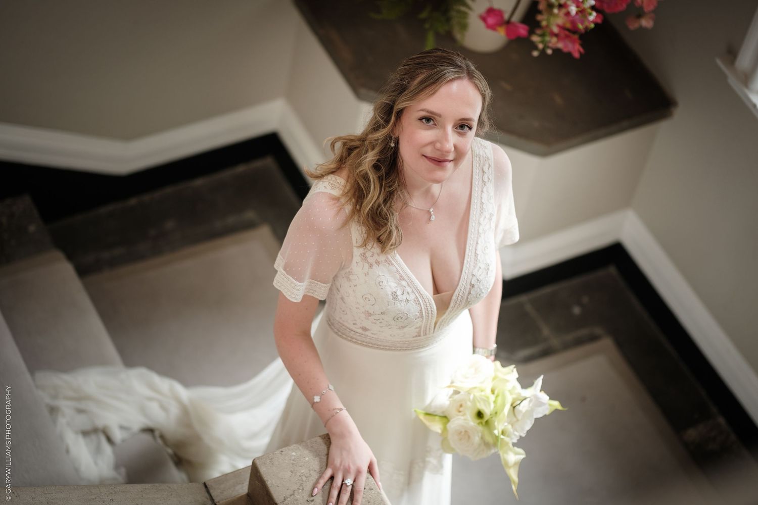 Bride in white lace gown holds bouquet on staircase, smiling down at camera in elegant venue.