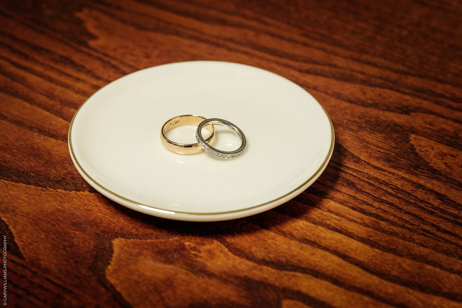 Two wedding rings, one gold and one diamond-encrusted, resting on a small white ceramic dish on a wooden surface.