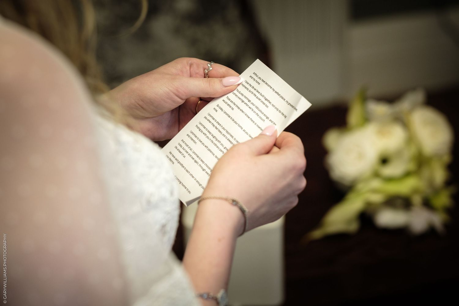 Bride in white lace dress reading wedding vows on a small card, with a floral bouquet blurred in the background.