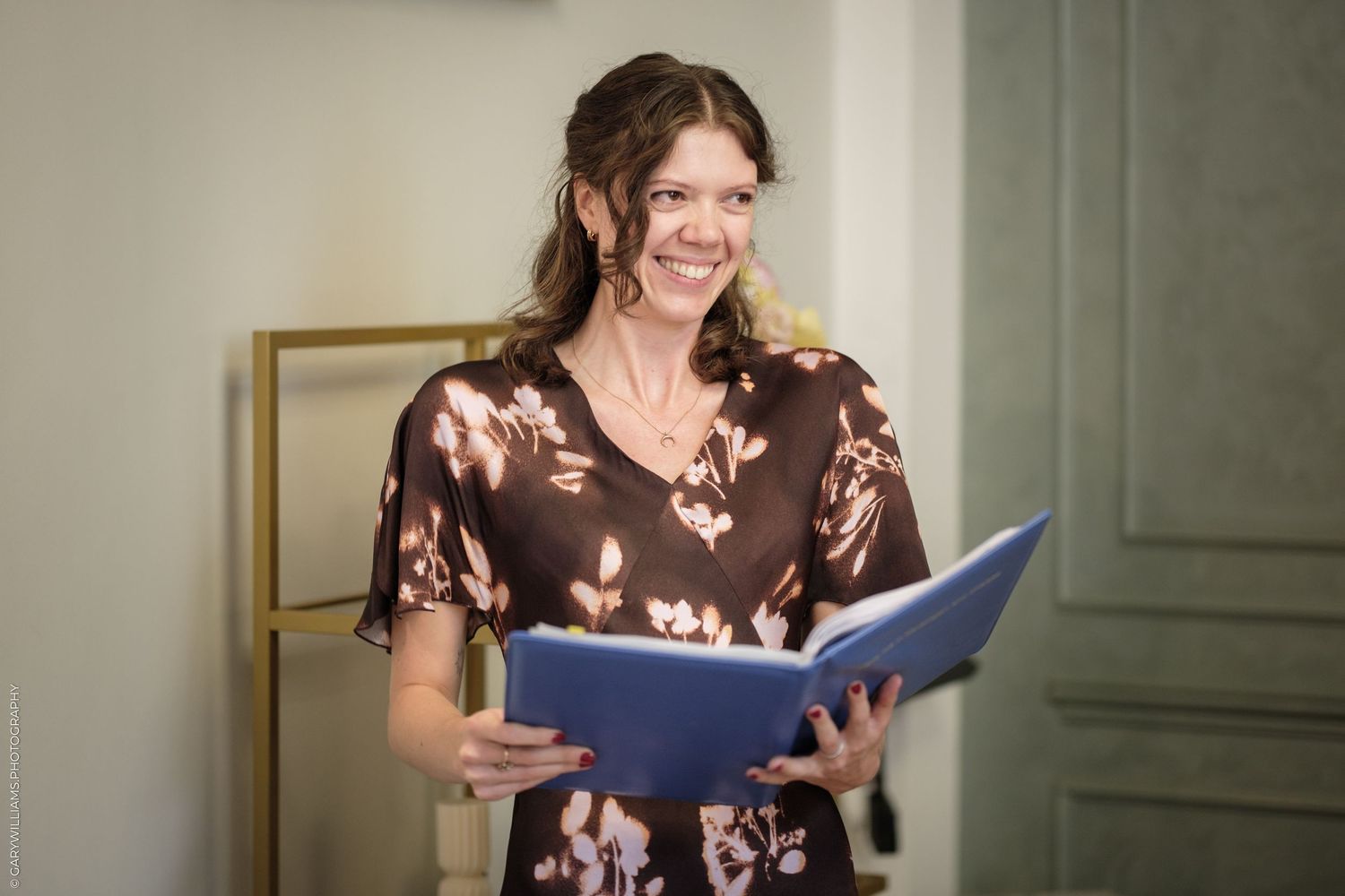 Smiling woman in floral dress holding a blue folder, standing in a bright elegant room.