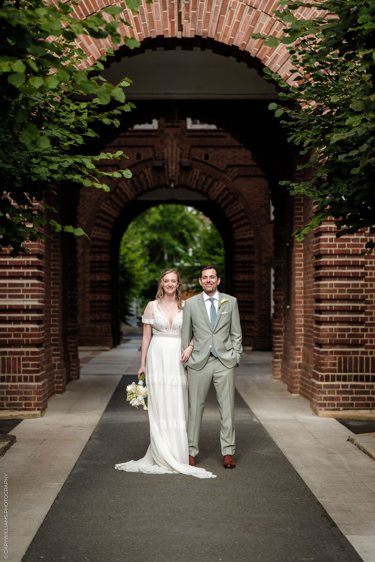 Bride and groom pose under brick archway draped in greenery, bride holding white bouquet in elegant white gown.