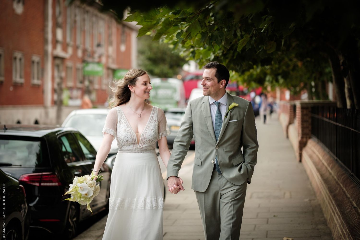 A bride and groom walk hand-in-hand on a London sidewalk, smiling at each other in elegant wedding attire.