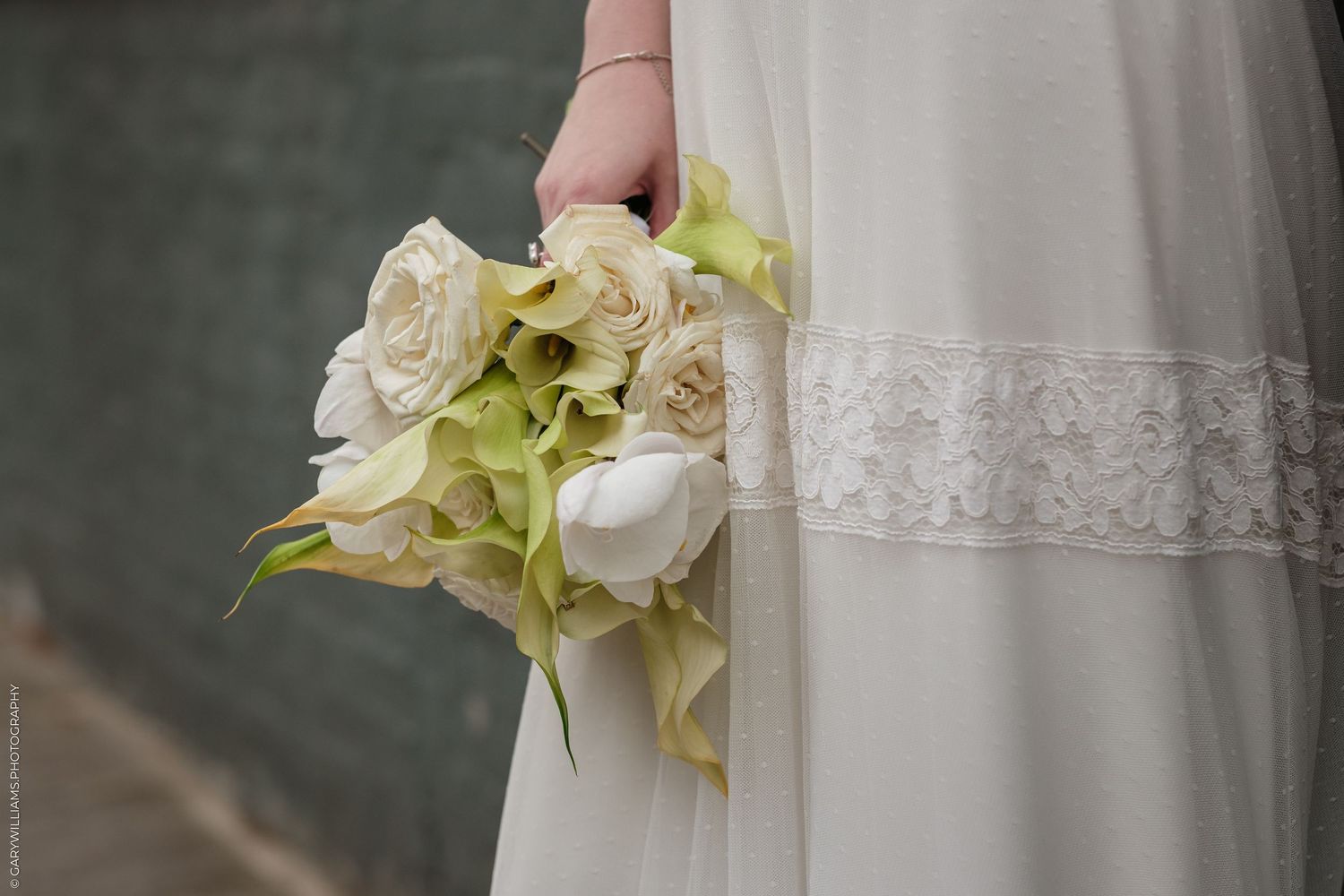 Bride in white lace dress holding elegant bouquet of cream roses and green calla lilies at wedding ceremony.