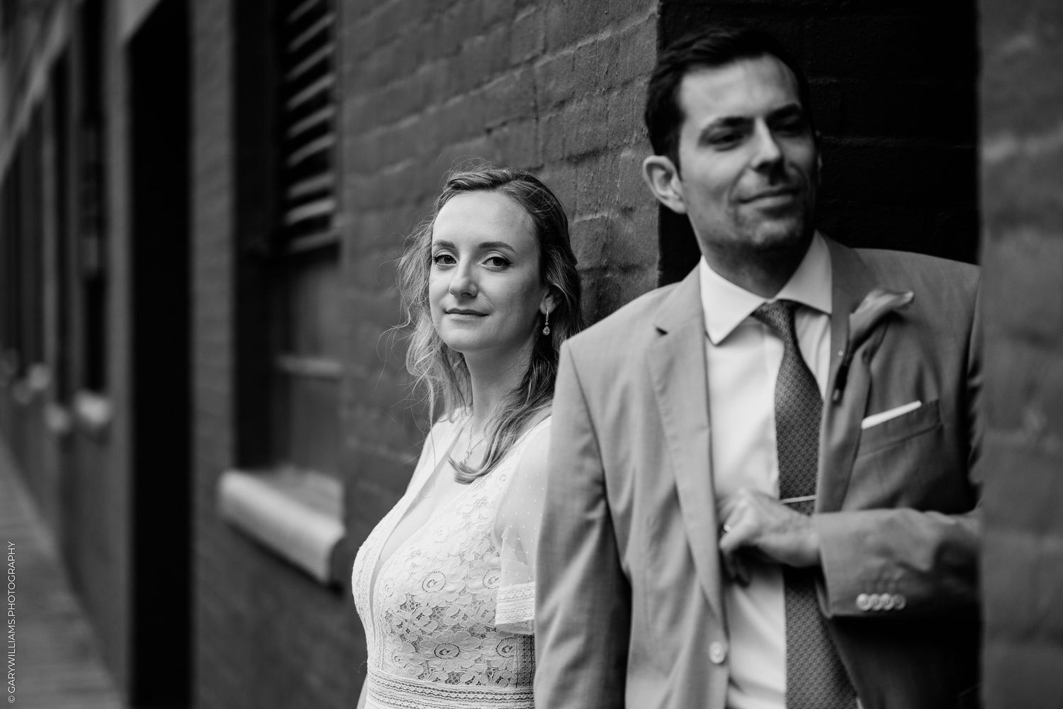 Black and white wedding photo of bride and groom posing against brick wall in elegant attire.