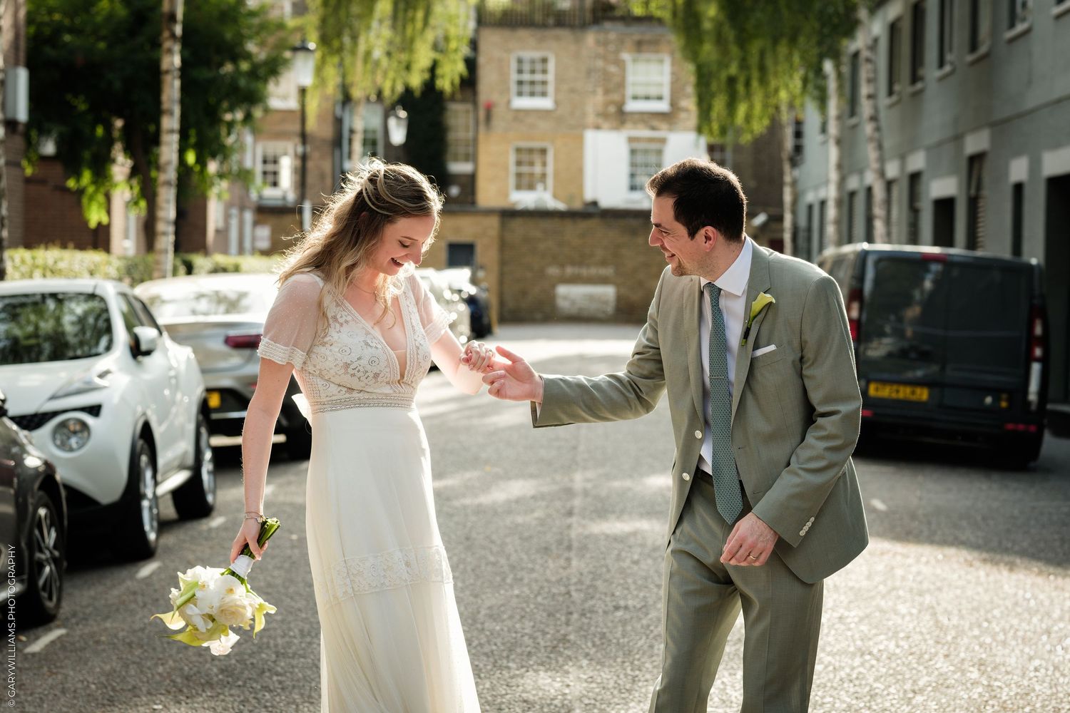 A bride and groom share a joyful moment on a sunny street, she holds white flowers as he reaches for her hand.