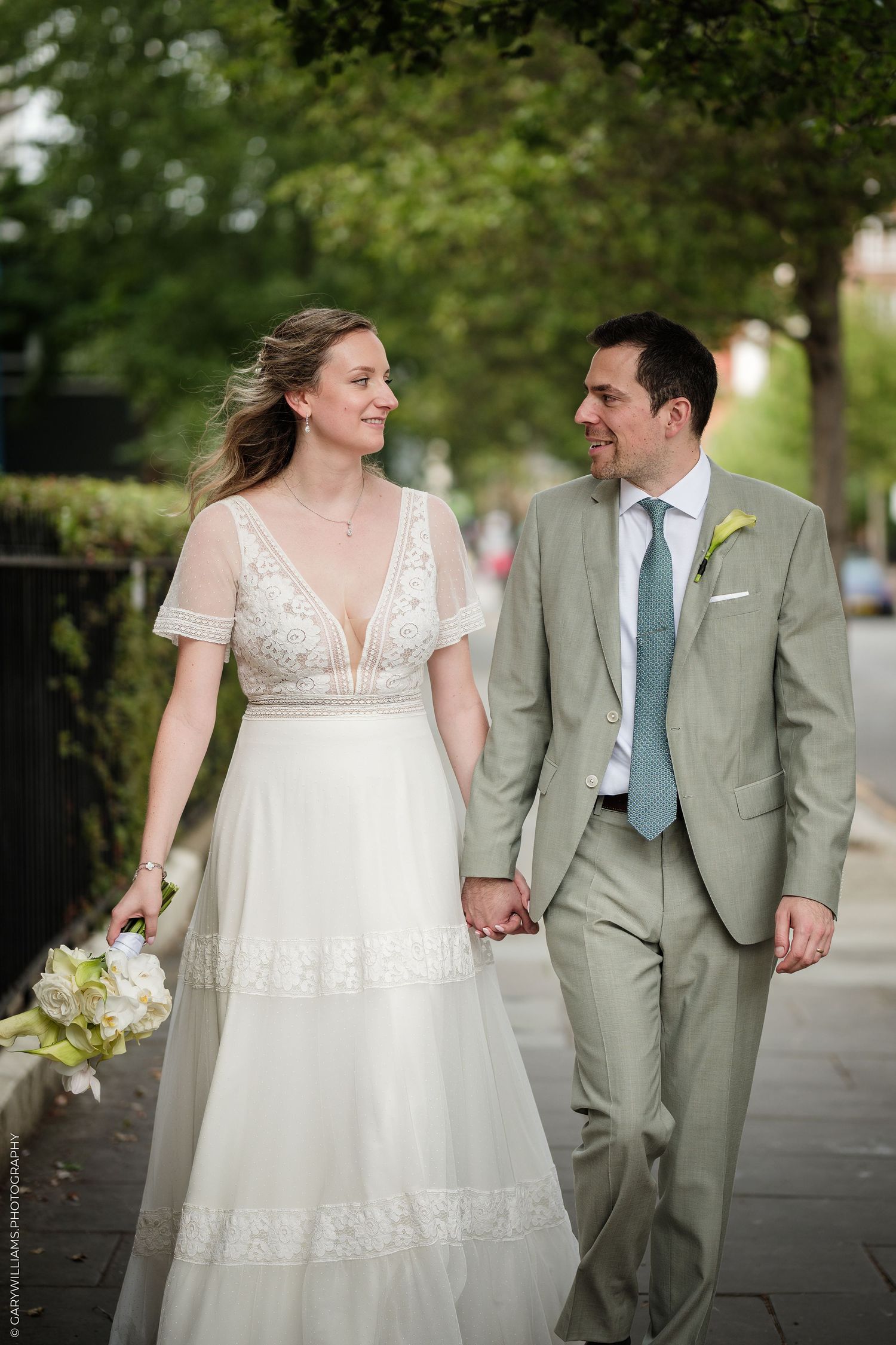 Bride and groom walk hand-in-hand on a tree-lined sidewalk, she in a white gown, he in a gray suit with blue tie.