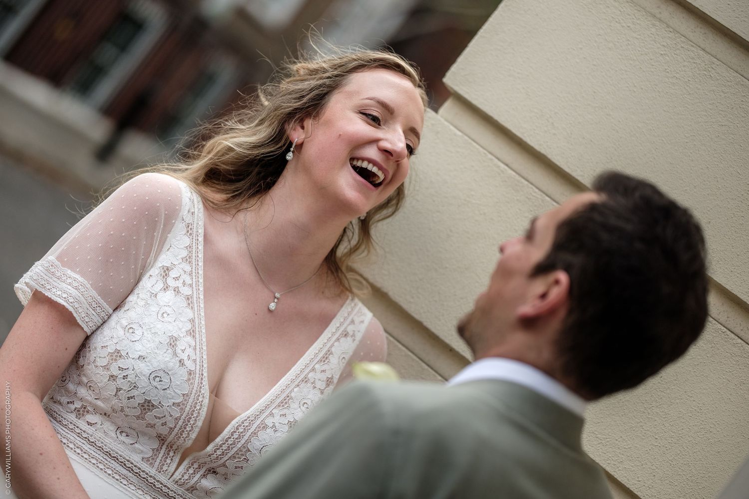 Bride in white lace dress laughing joyfully with groom in suit during outdoor wedding photo session.