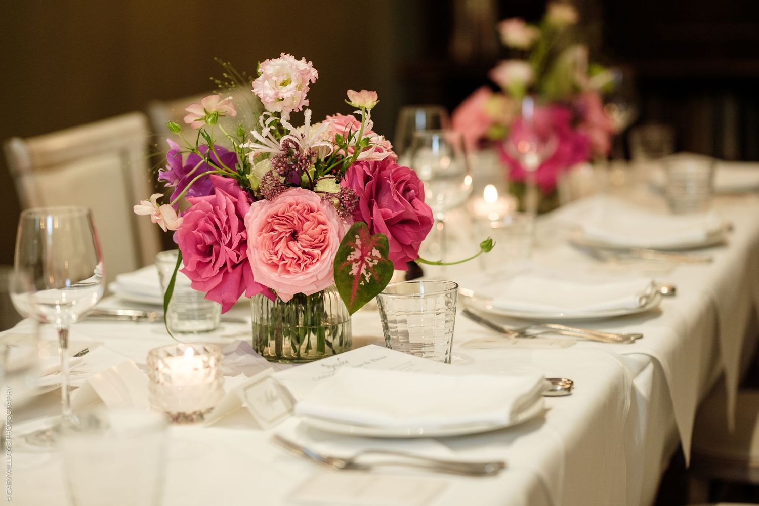 Elegant wedding reception table with pink and purple floral centerpiece, white linens, fine china, and crystal glassware.