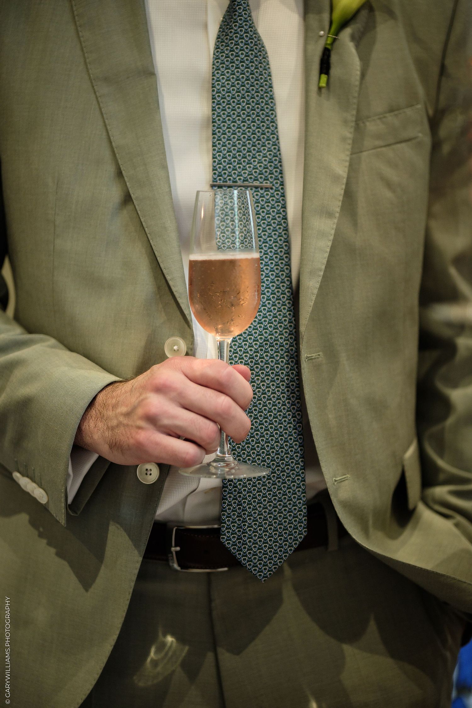 Man in gray suit holding a champagne flute with rose sparkling wine, wearing a teal patterned tie.
