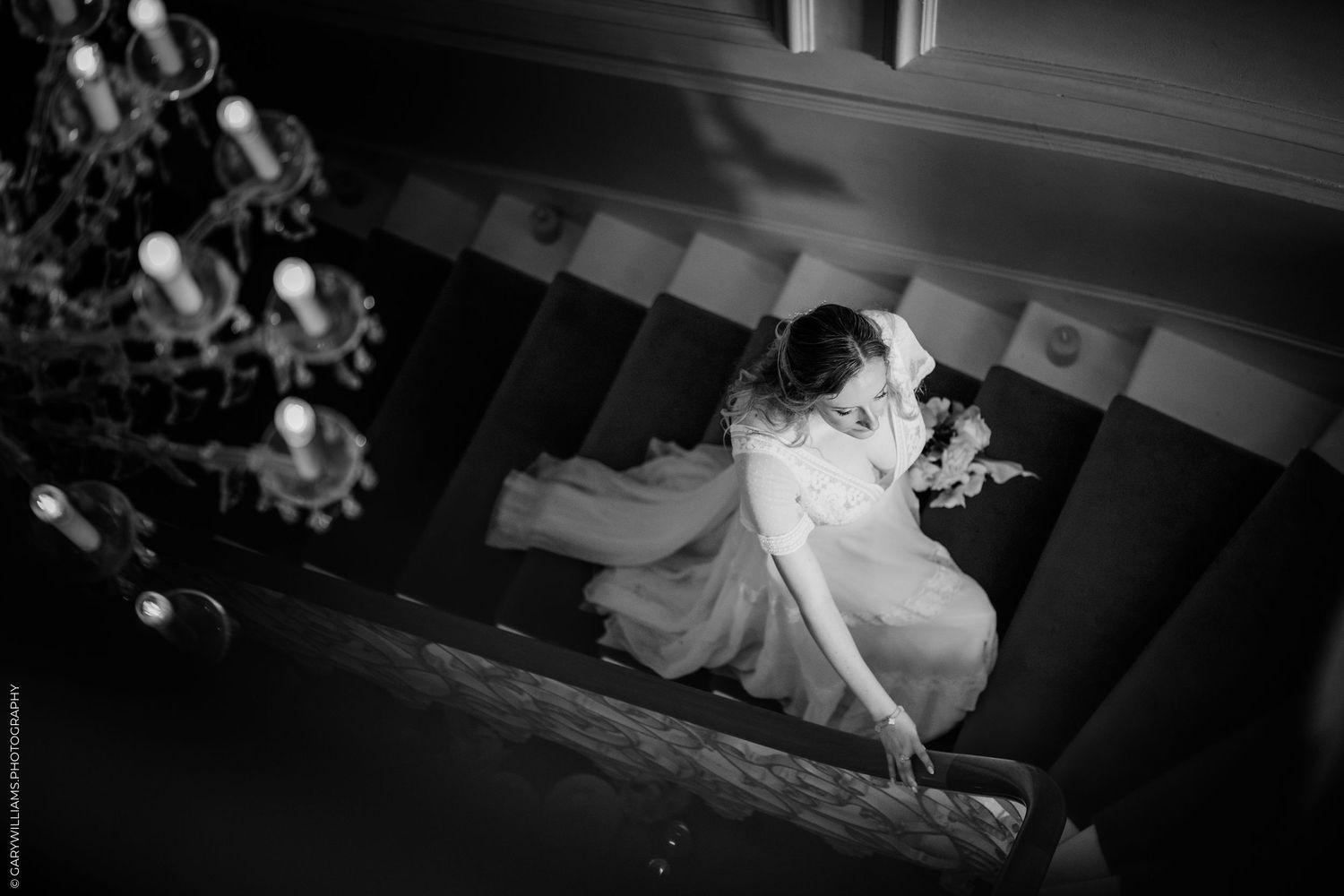 Black and white wedding photo of bride descending elegant staircase with chandelier, captured from above.