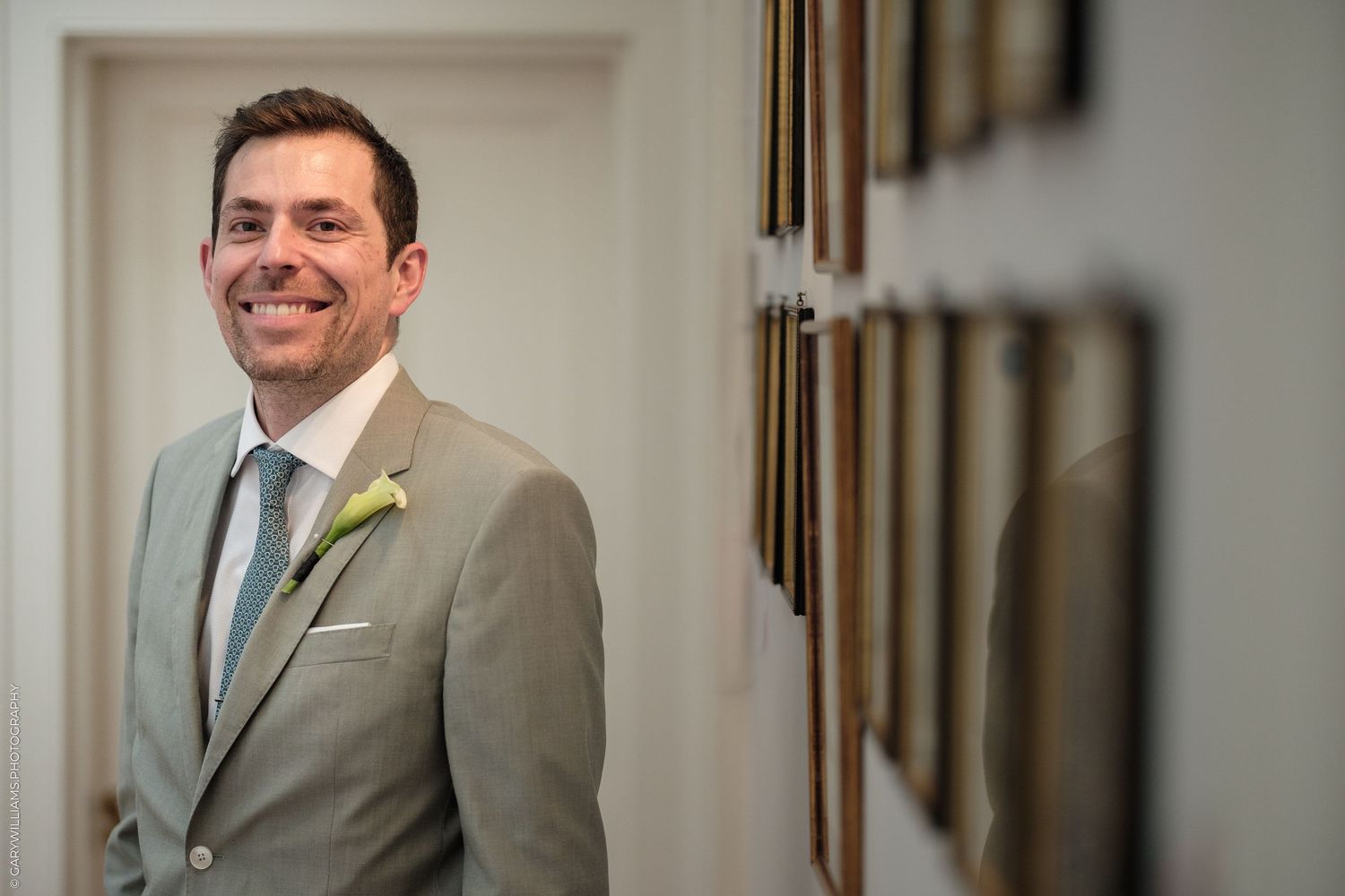 Smiling groom in light gray suit with boutonniere standing in elegant hallway with framed pictures.