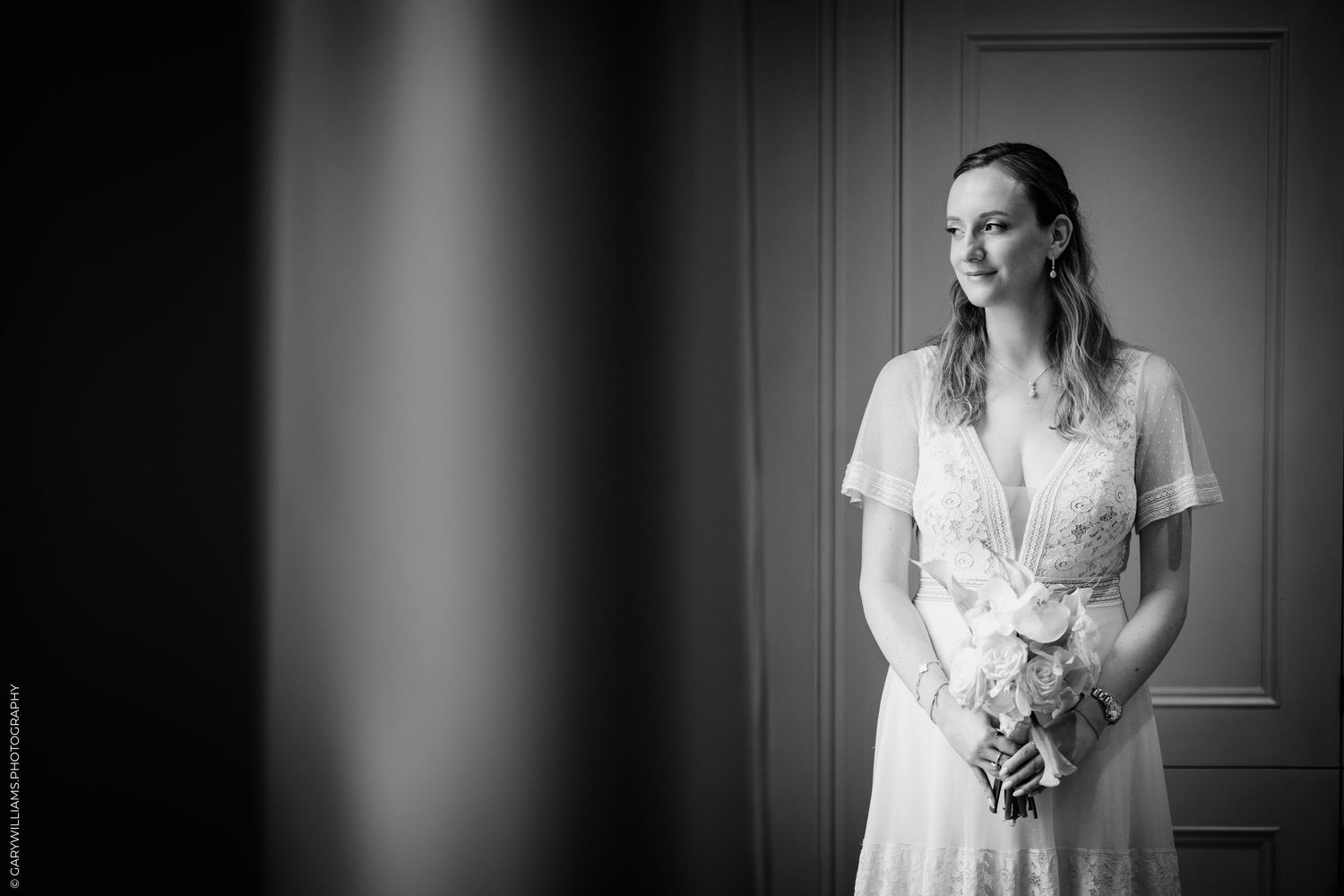 Black and white portrait of a bride in a lace wedding dress holding a bouquet, standing elegantly in soft natural light.