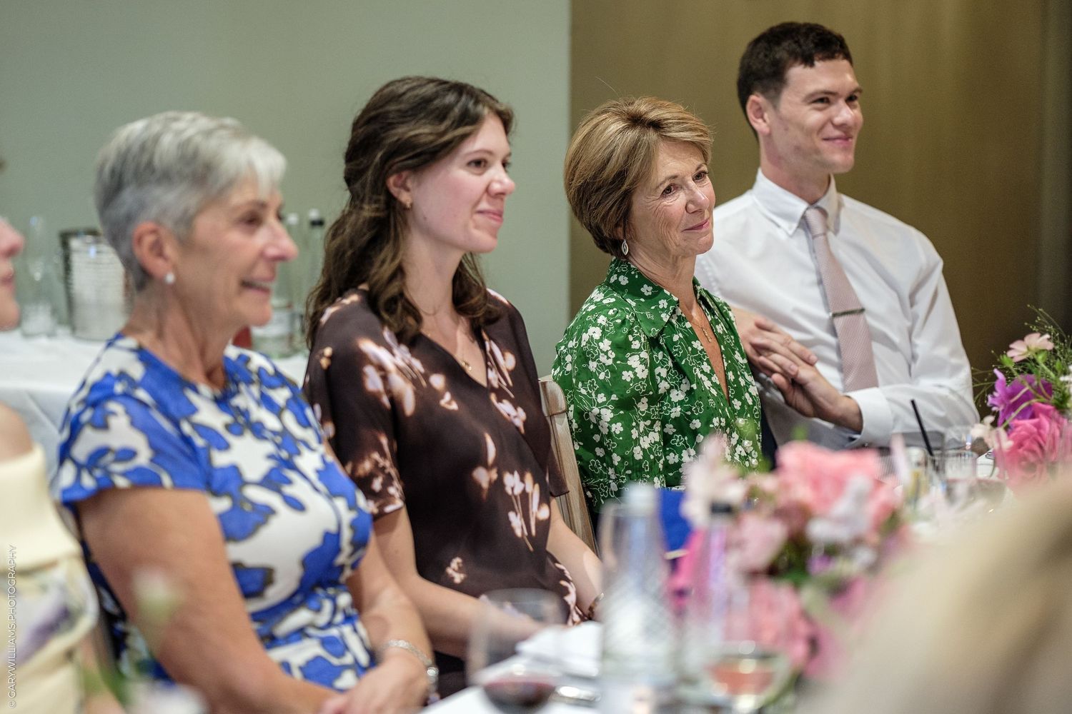 Four guests smile and applaud at a formal event, seated at a floral-decorated banquet table indoors.