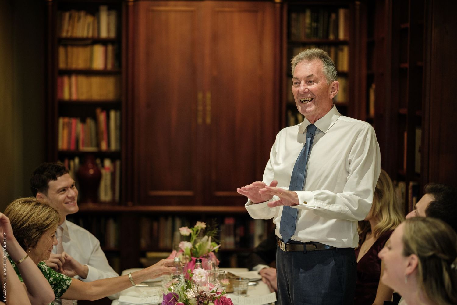 Man in shirt and tie smiling while giving a speech at an elegant dinner party with guests seated around a decorated table.