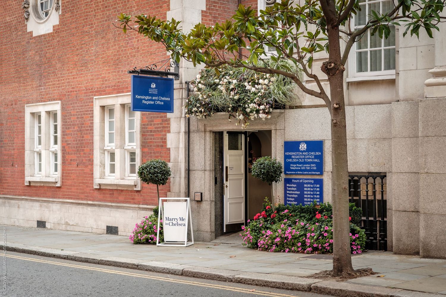 Brick building entrance with blue signage, flowering plants, and a tree-lined cobblestone street in London.