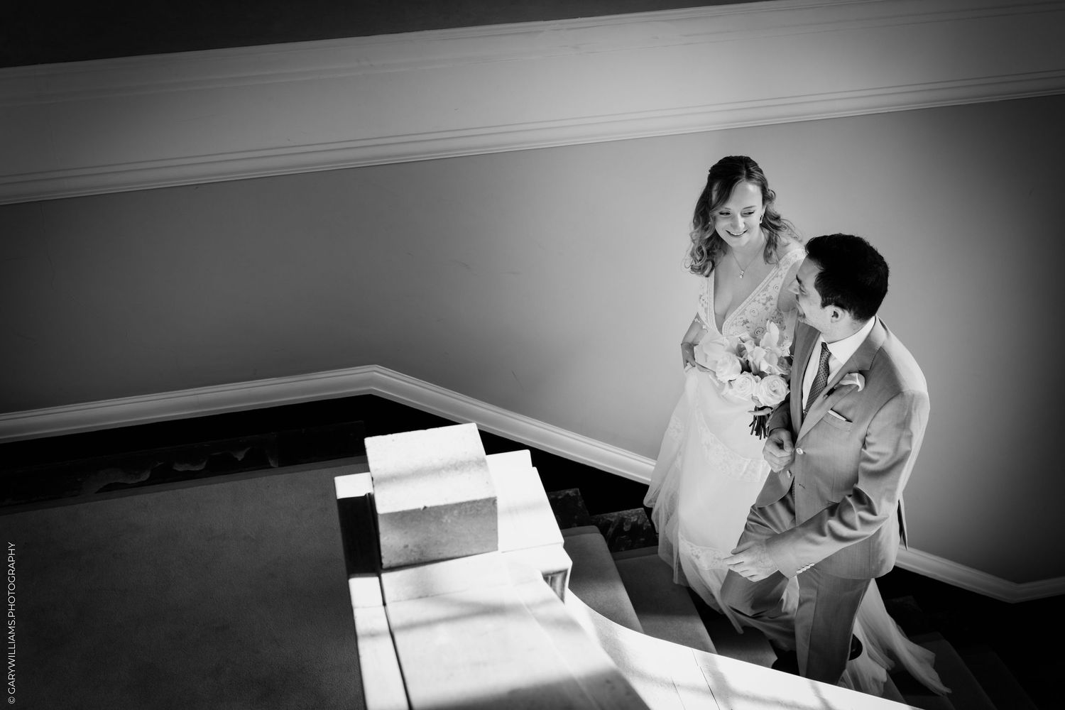 Black and white photo of a groom kneeling before his bride in a wedding dress, holding a bouquet indoors.