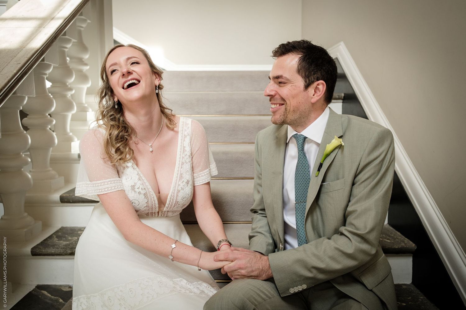 Happy bride and groom laughing together on staircase, bride in white lace dress, groom in tan suit with green tie.
