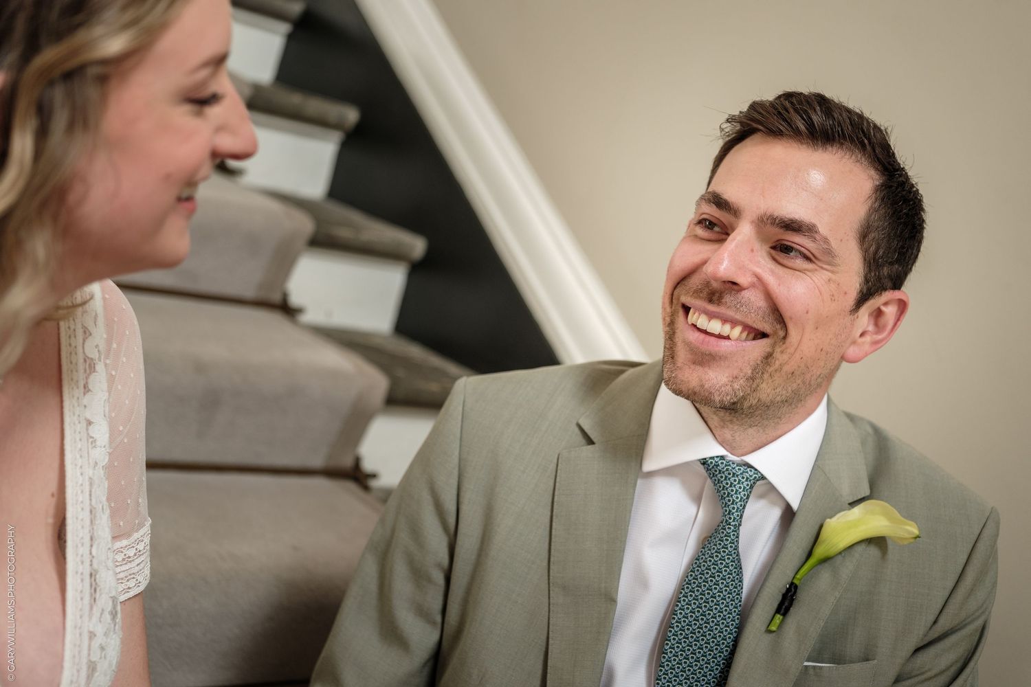 Groom in sage suit smiling at bride during wedding ceremony near staircase.
