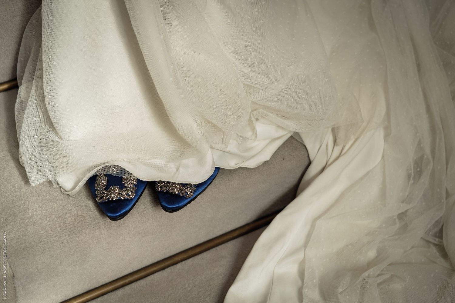Blue rhinestone bridal shoes peeking beneath a flowing white wedding dress on carpeted stairs.
