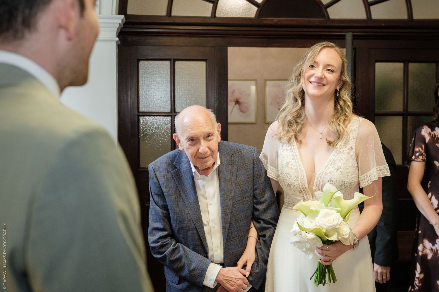 Bride in white gown holding bouquet smiles at groom during indoor wedding ceremony with elderly guest nearby.
