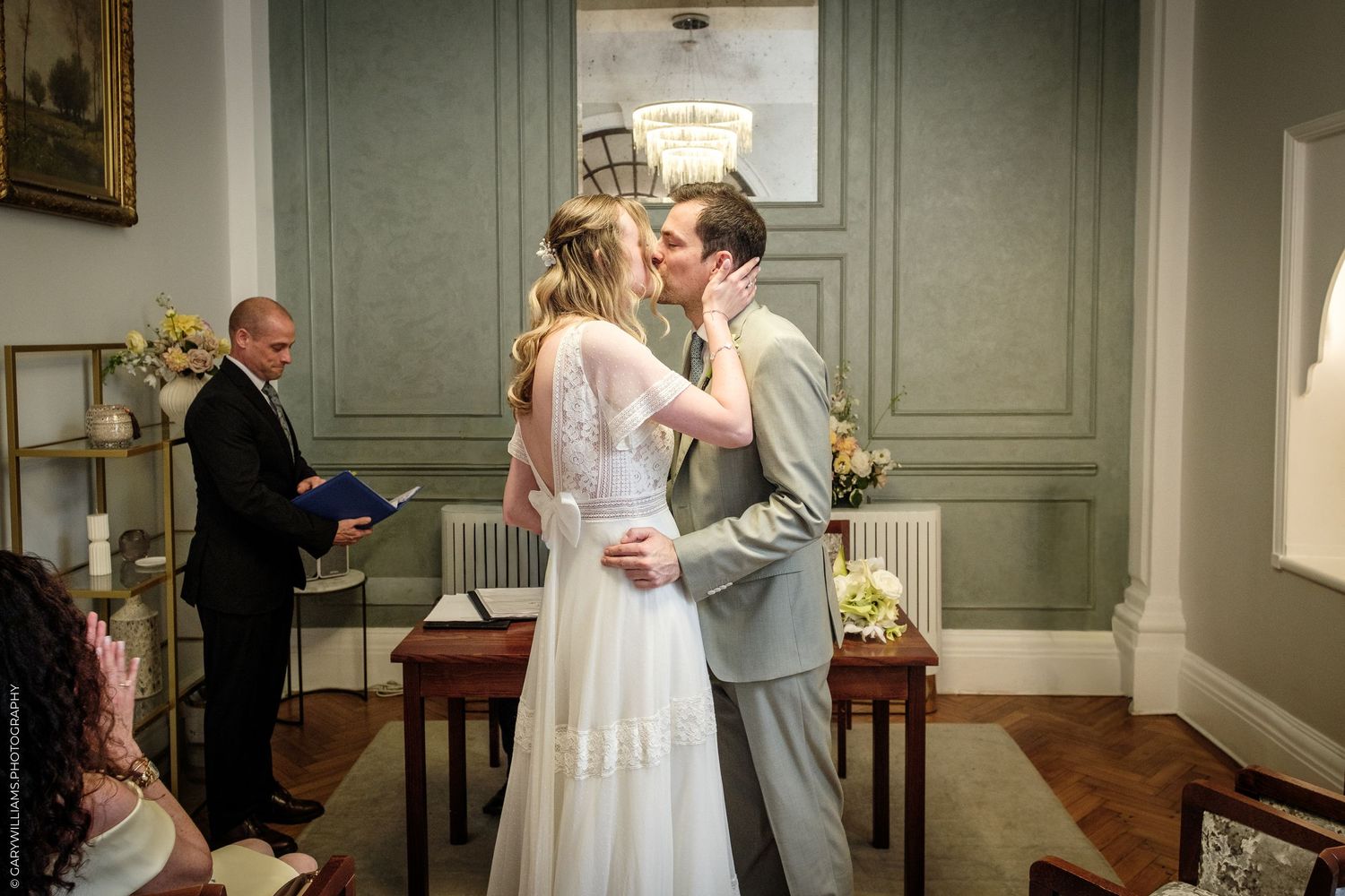 Bride and groom share first kiss at wedding ceremony while officiant reads from book in elegant gray room.