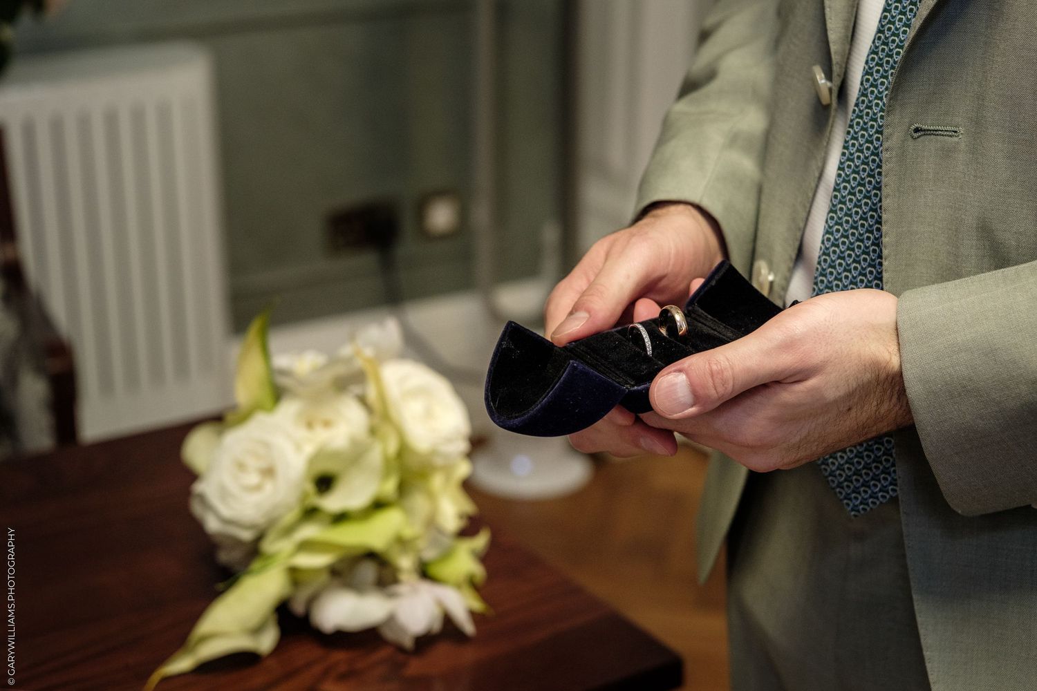 Groom in sage suit holds wedding rings in black box, with white floral bouquet on table in background.