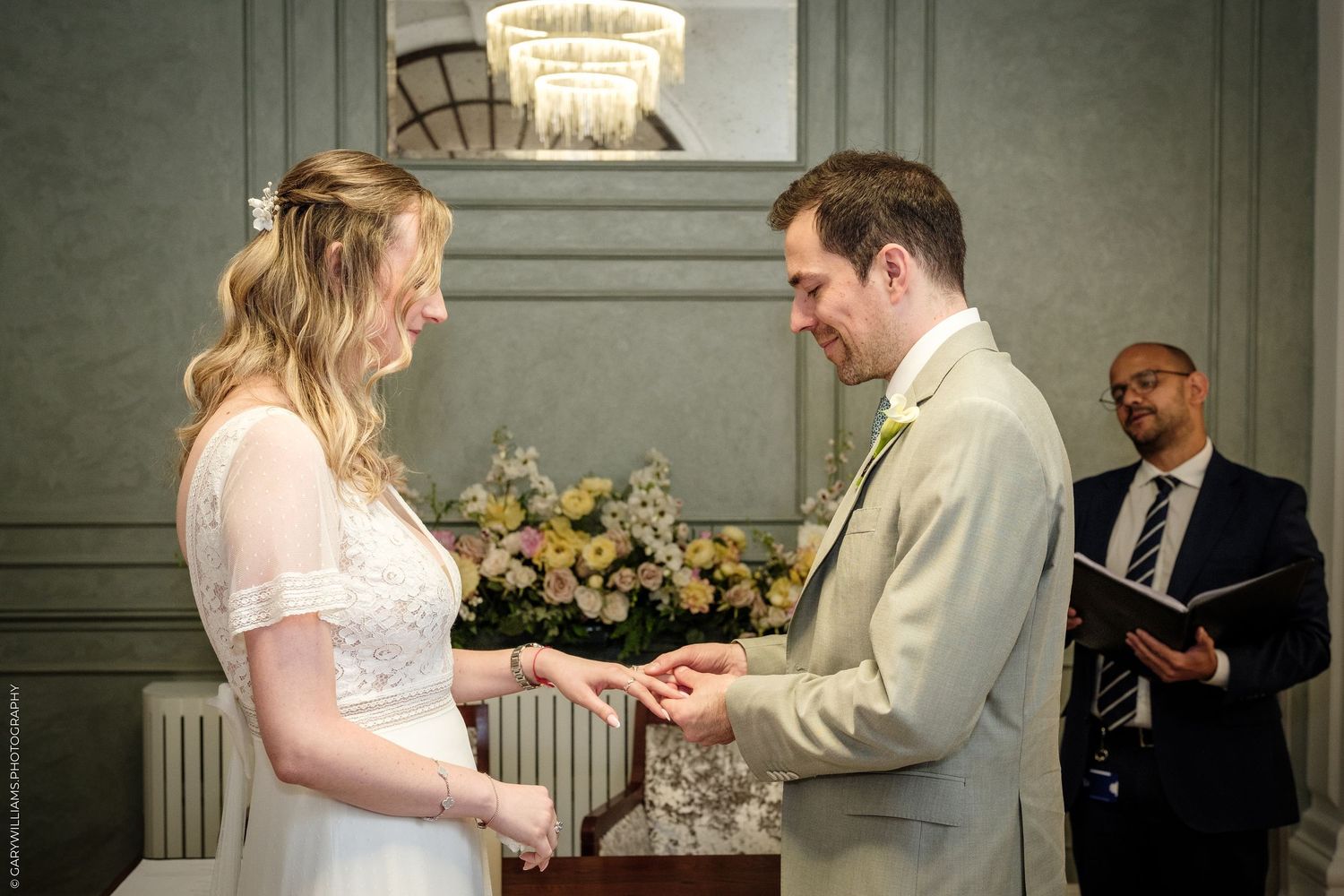 Bride and groom exchange rings during wedding ceremony, officiant holding book in background.