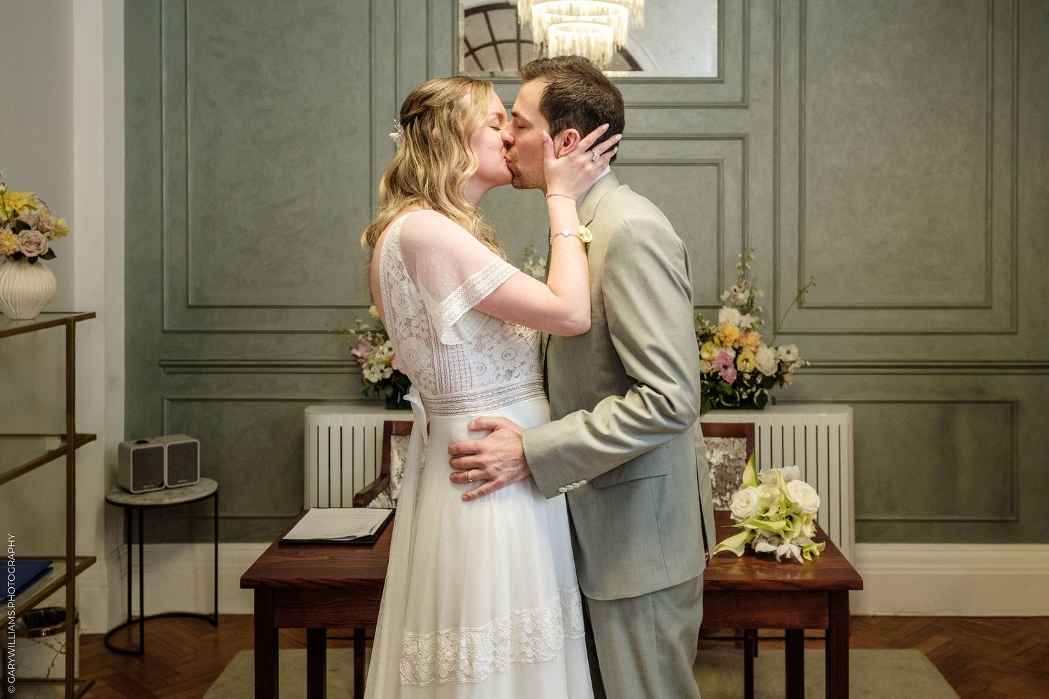 A bride and groom share their first kiss at a wedding ceremony in an elegant room with green walls.
