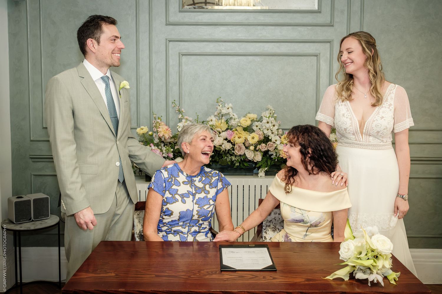 Wedding party laughing at signing table with bride, groom, and guests in elegant blue-gray room with floral arrangement.
