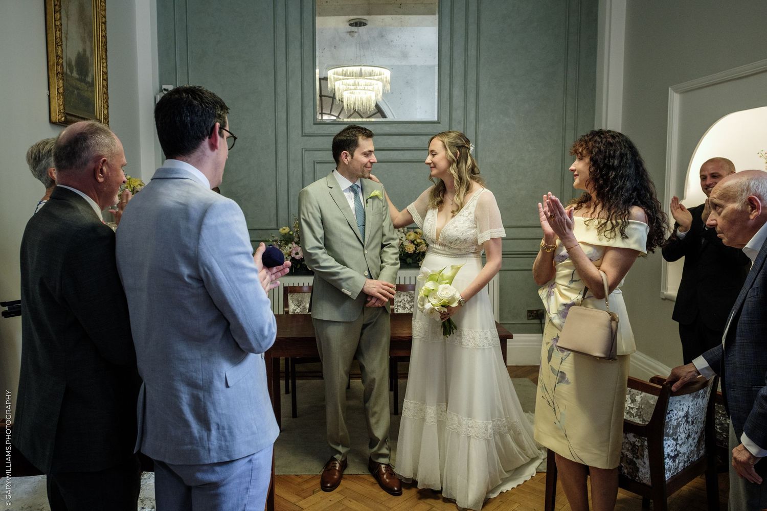 Bride and groom exchanging vows in an elegant indoor wedding ceremony surrounded by smiling guests clapping.