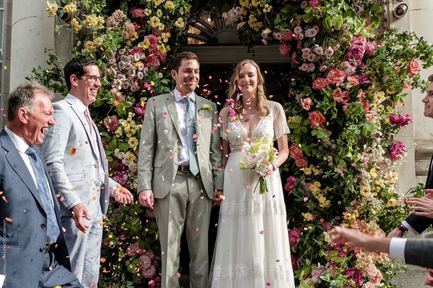Bride and groom smile as guests throw confetti outside a floral arch entrance on their wedding day.