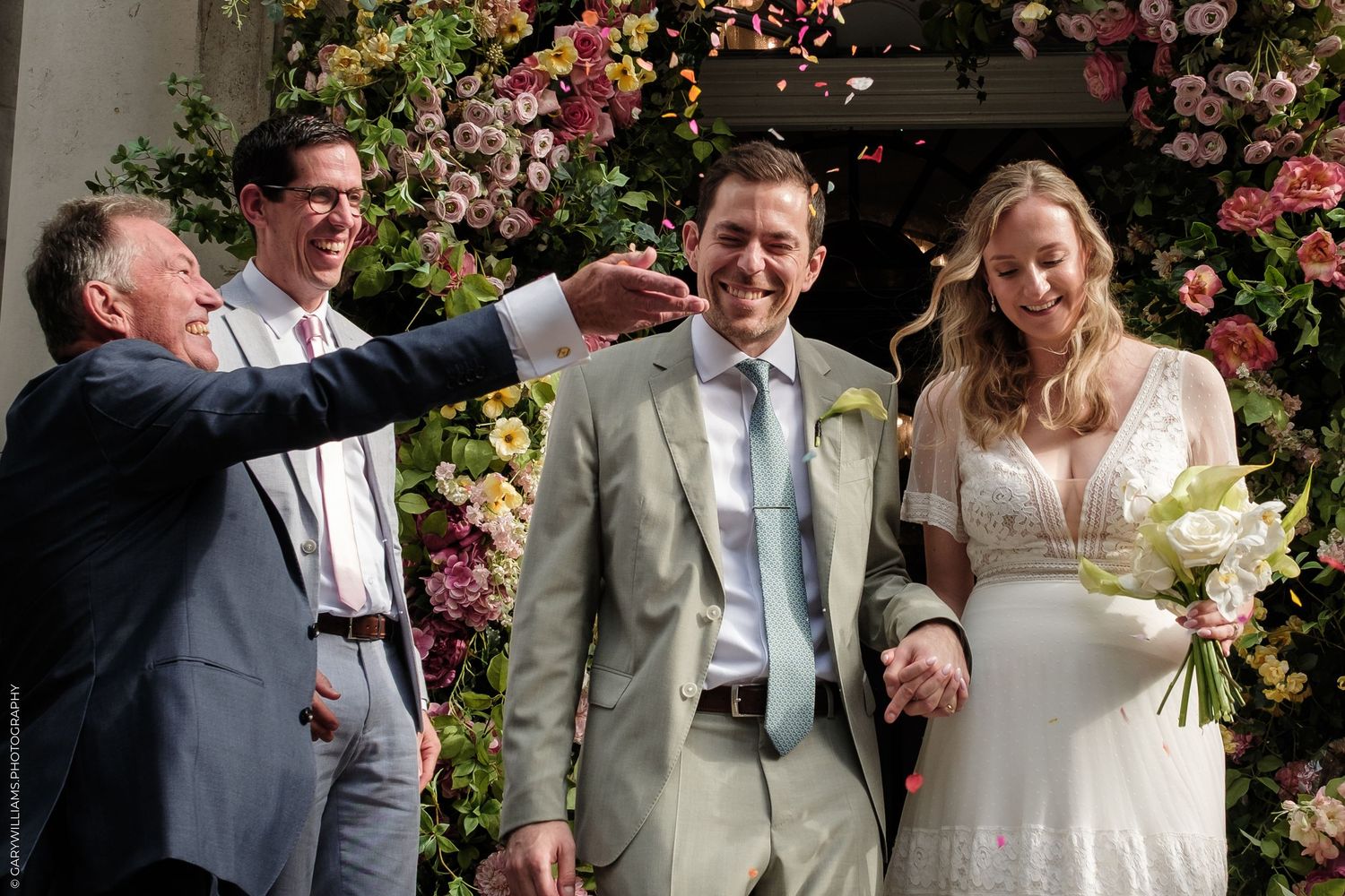 Joyful wedding ceremony with bride and groom laughing as guests celebrate before a lush floral backdrop.