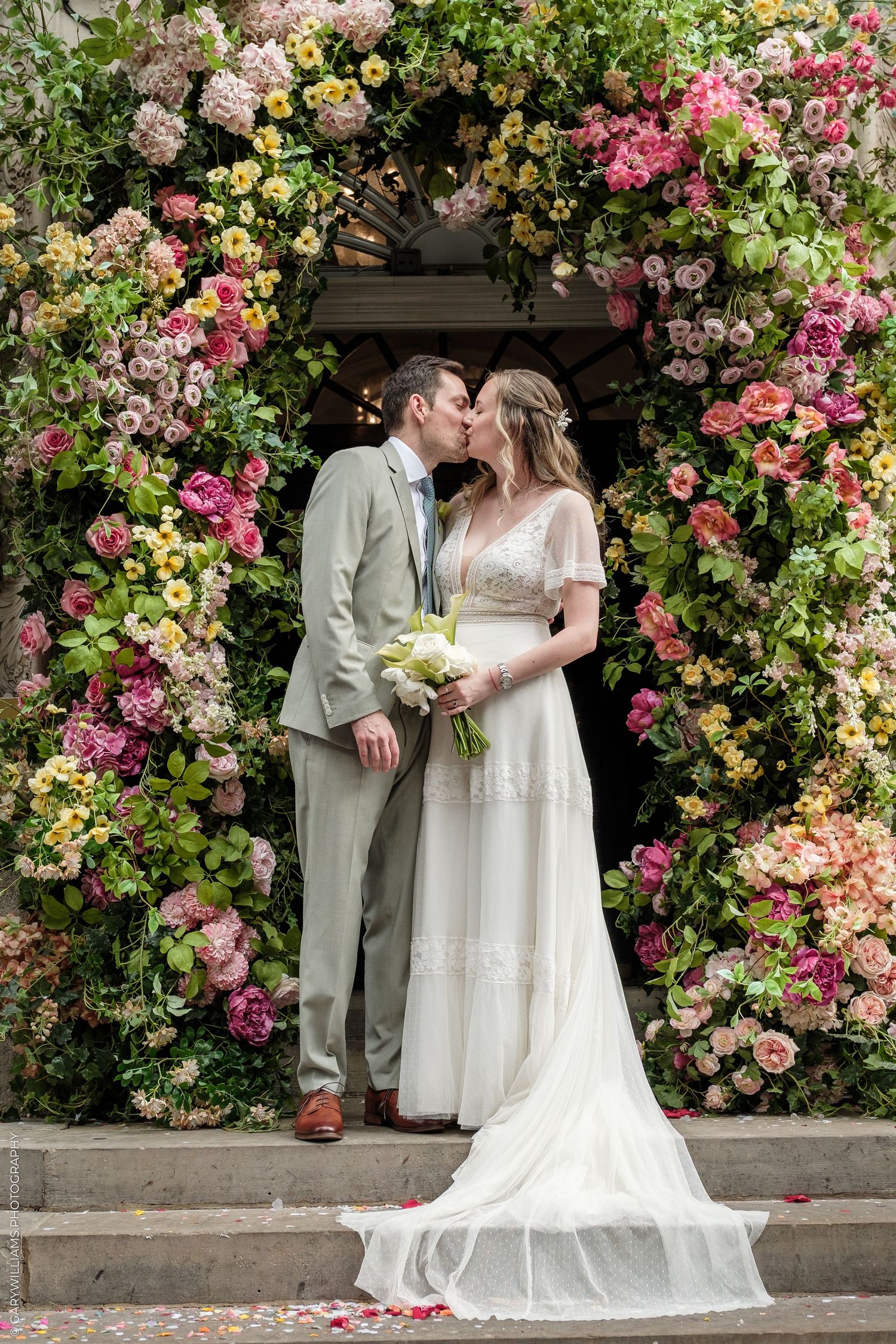 Bride and groom kissing under a stunning floral arch with pink, yellow, and white blooms at their wedding ceremony.