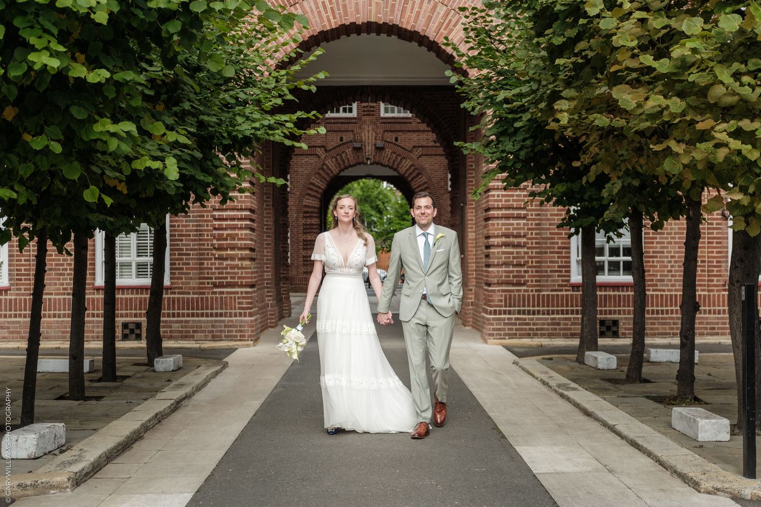 Bride and groom walking hand in hand along a tree-lined path before a brick archway on their wedding day.