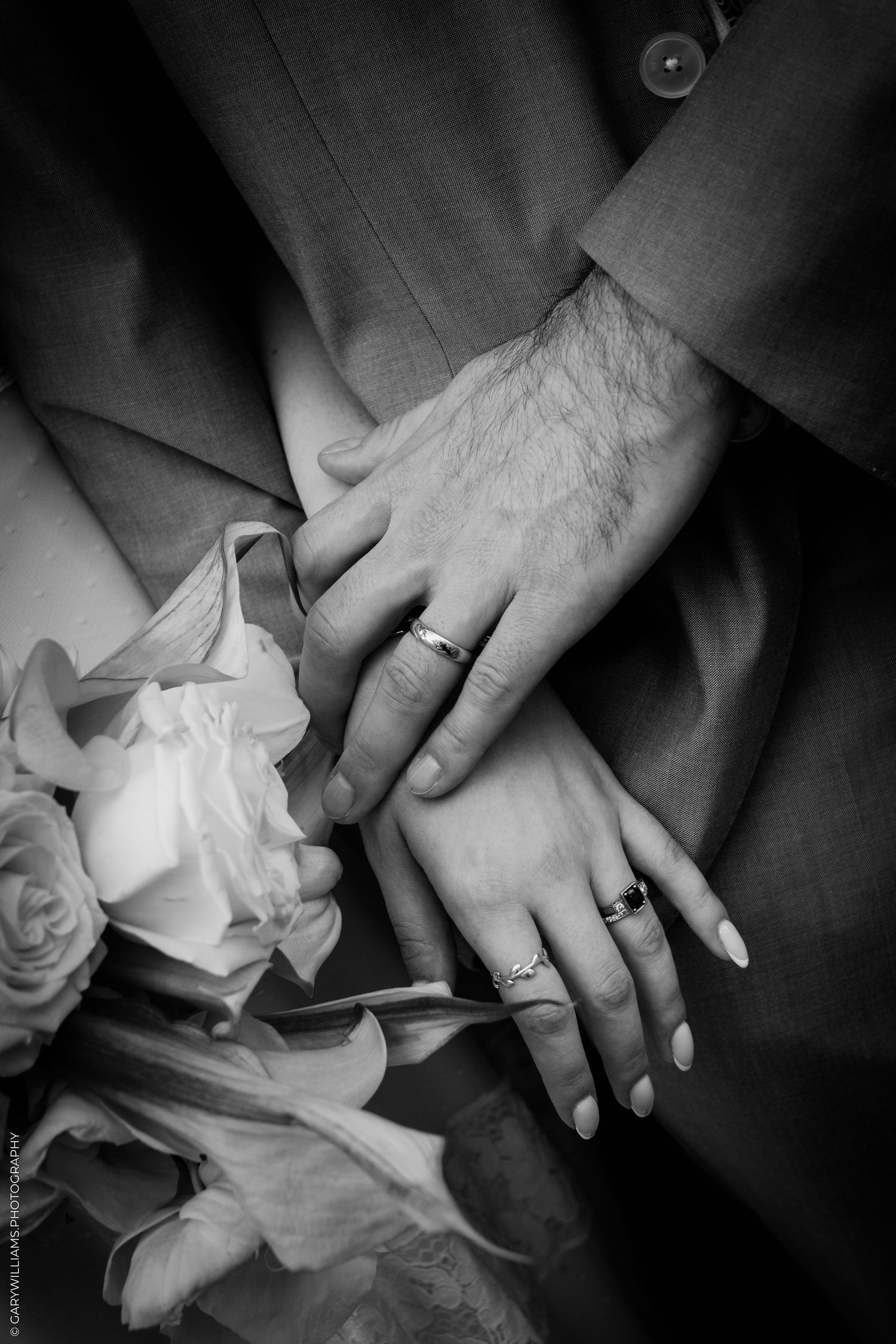 Black and white photo of a bride and groom holding hands, displaying wedding rings, with a bouquet of roses nearby.