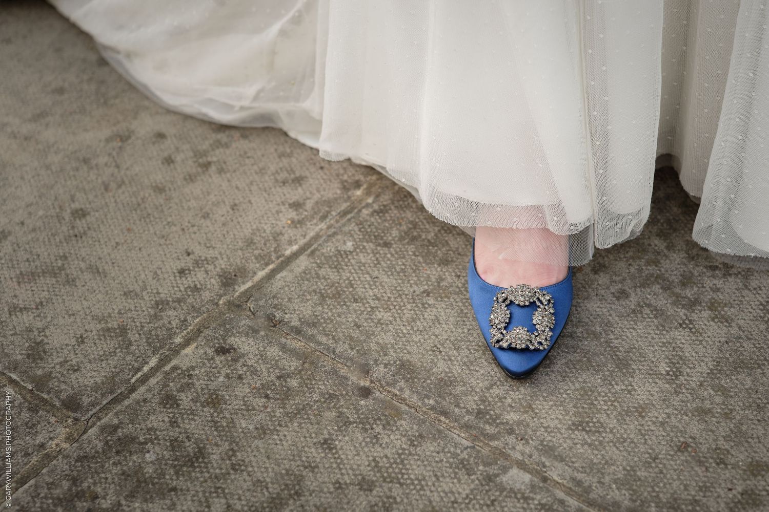 Bride wearing a white tulle gown and blue Manolo Blahnik heels with jeweled buckle on concrete floor.