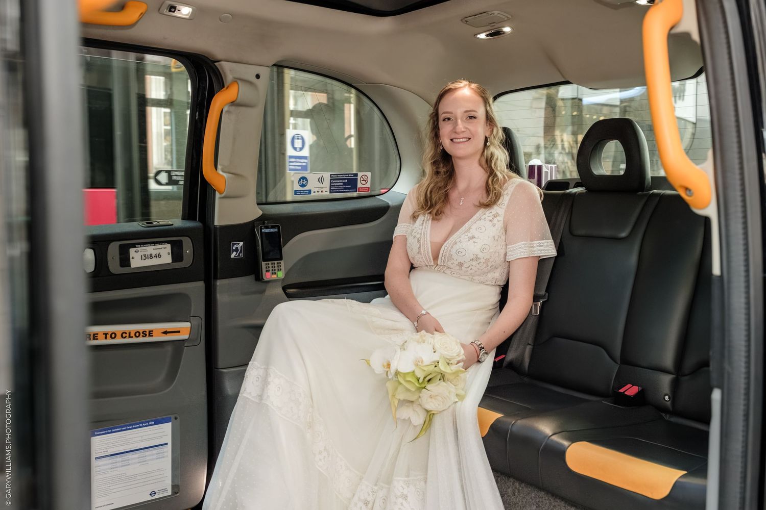 Bride in white wedding dress holding bouquet sits in black London taxi cab interior.
