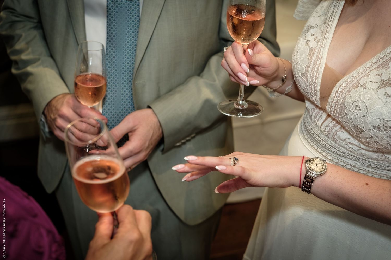 Wedding guests clinking champagne flutes during a toast, bride in lace gown visible with gold watch and ring.