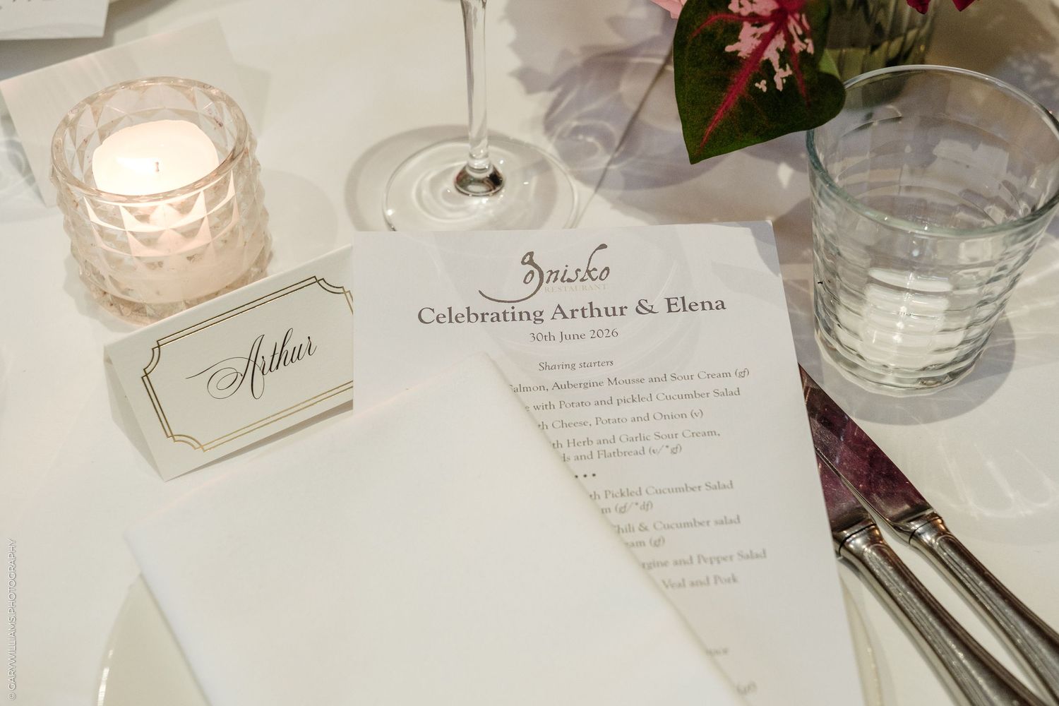 Elegant wedding reception table setting with Arthur place card, menu for Arthur & Elena celebration, candle, and glassware.