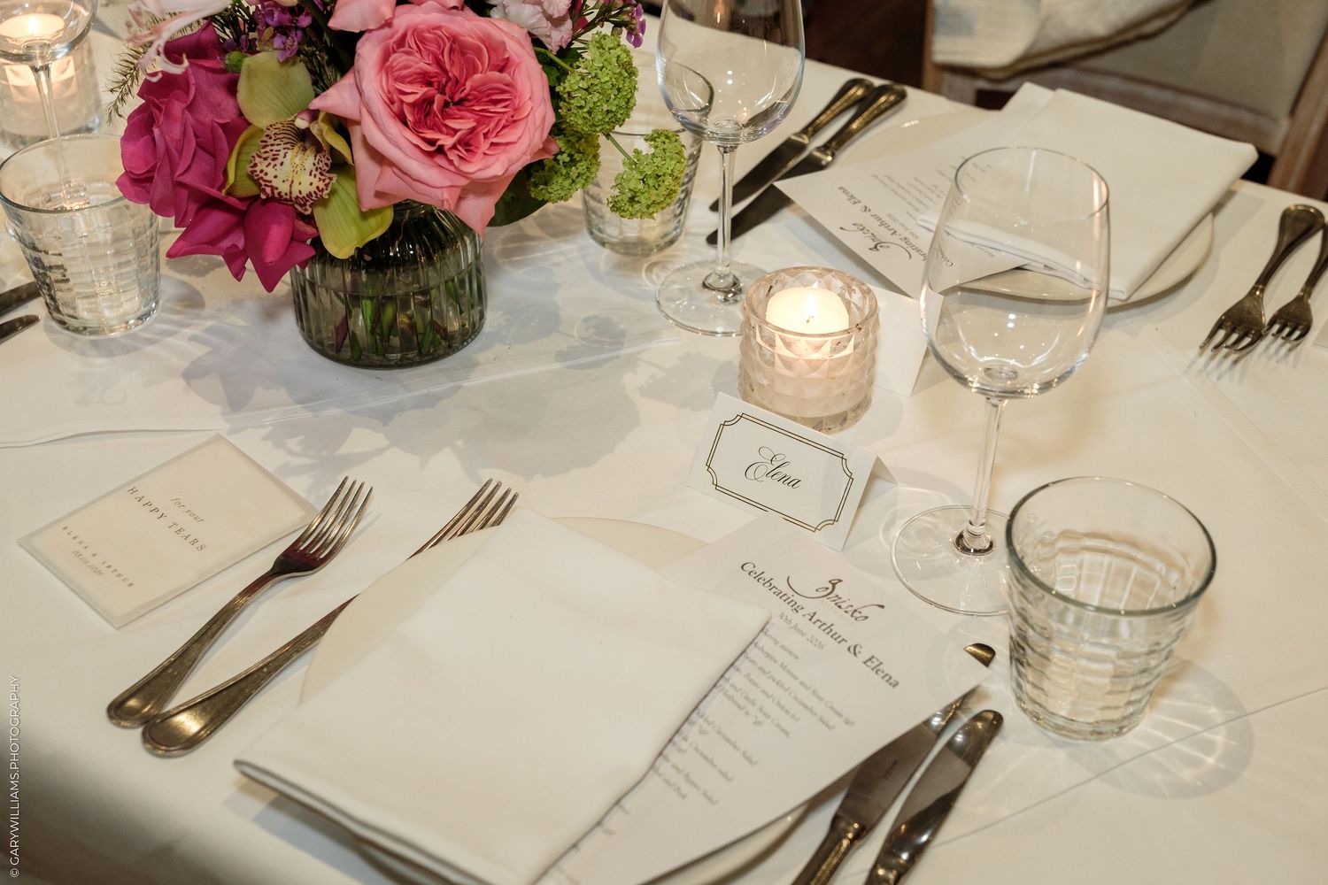 Elegant wedding reception table setting with pink floral centerpiece, white linens, crystal glassware, and candlelight.