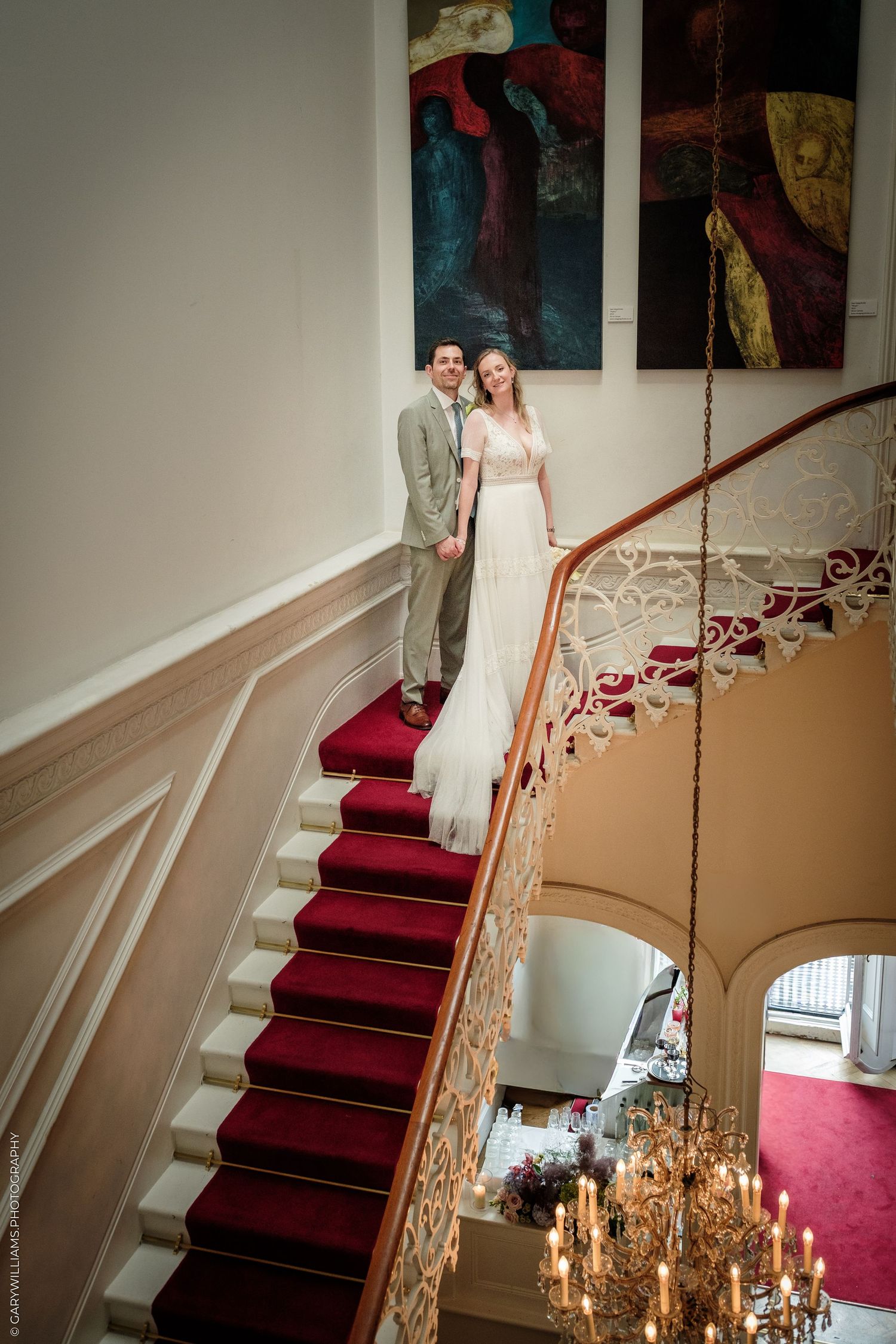 Bride and groom stand elegantly on a grand staircase with red carpet and ornate railing in a luxurious wedding venue.