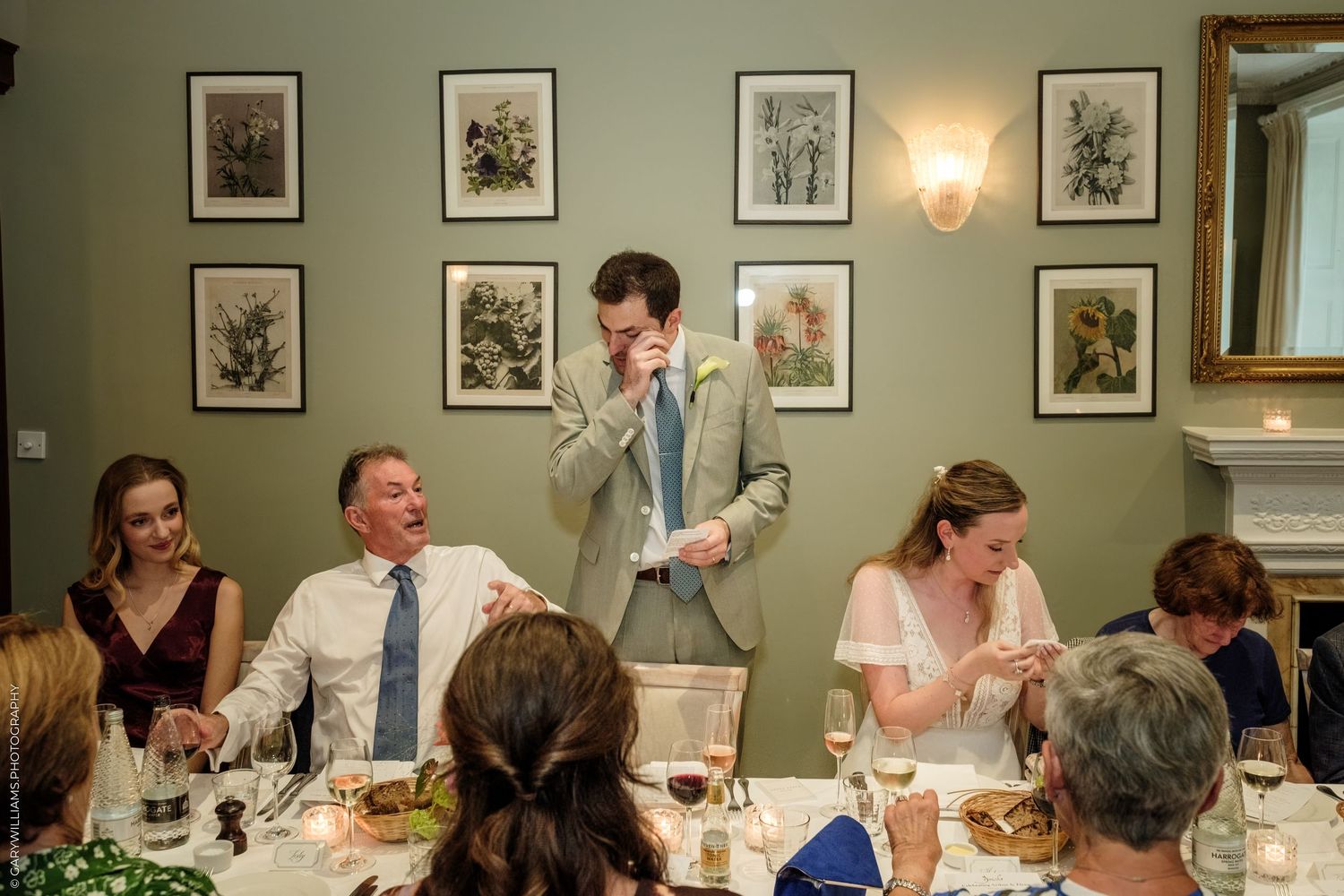 Emotional groom giving wedding speech at elegant dinner reception, guests laughing and smiling around decorated table.