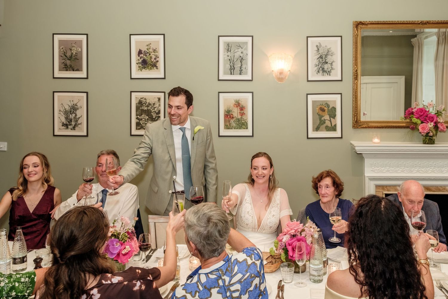 Bride and groom toast guests at elegant wedding reception with floral arrangements and botanical art on green walls.