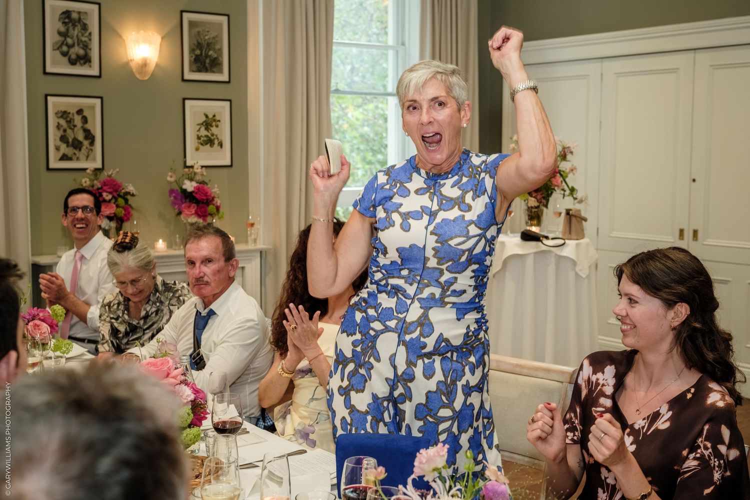 Woman in blue floral dress celebrating joyfully at an elegant wedding reception dinner party.