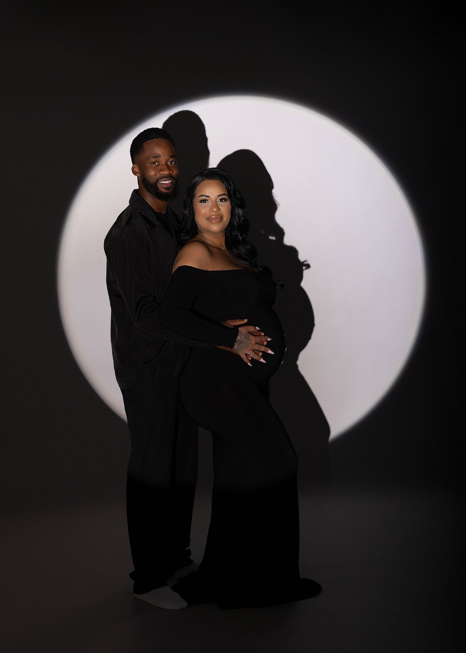 Couple in matching black outfits pose for maternity photo against dramatic circular white light on dark background.