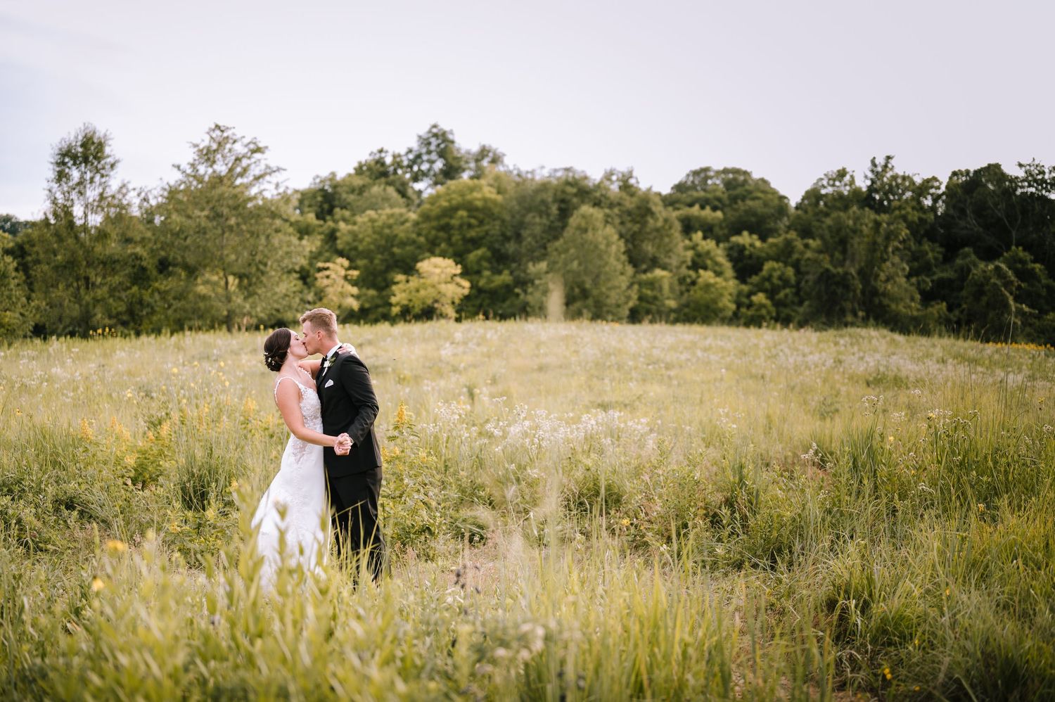 Bride and groom embracing in a lush green meadow with wildflowers and trees in the background on their wedding day.
