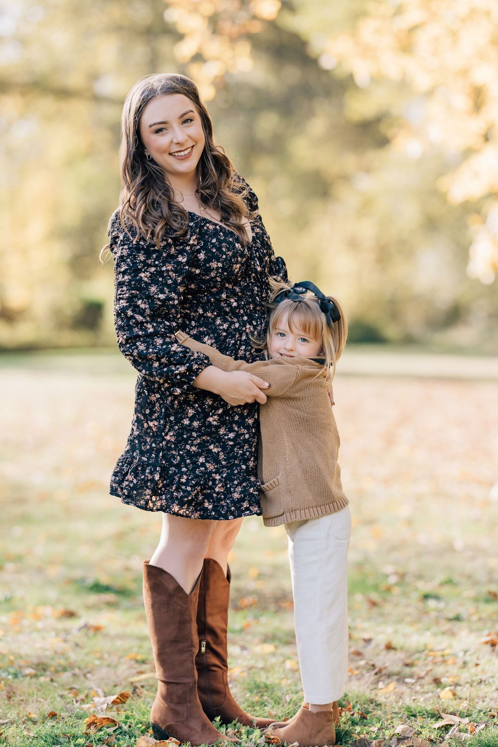 A smiling woman in a floral dress and brown boots hugs a young girl in a tan sweater outdoors in autumn.