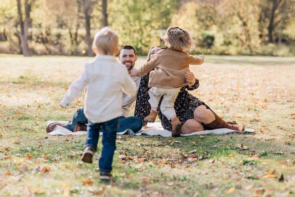 Family enjoying autumn park picnic, children running to parents sitting on blanket among fallen leaves.