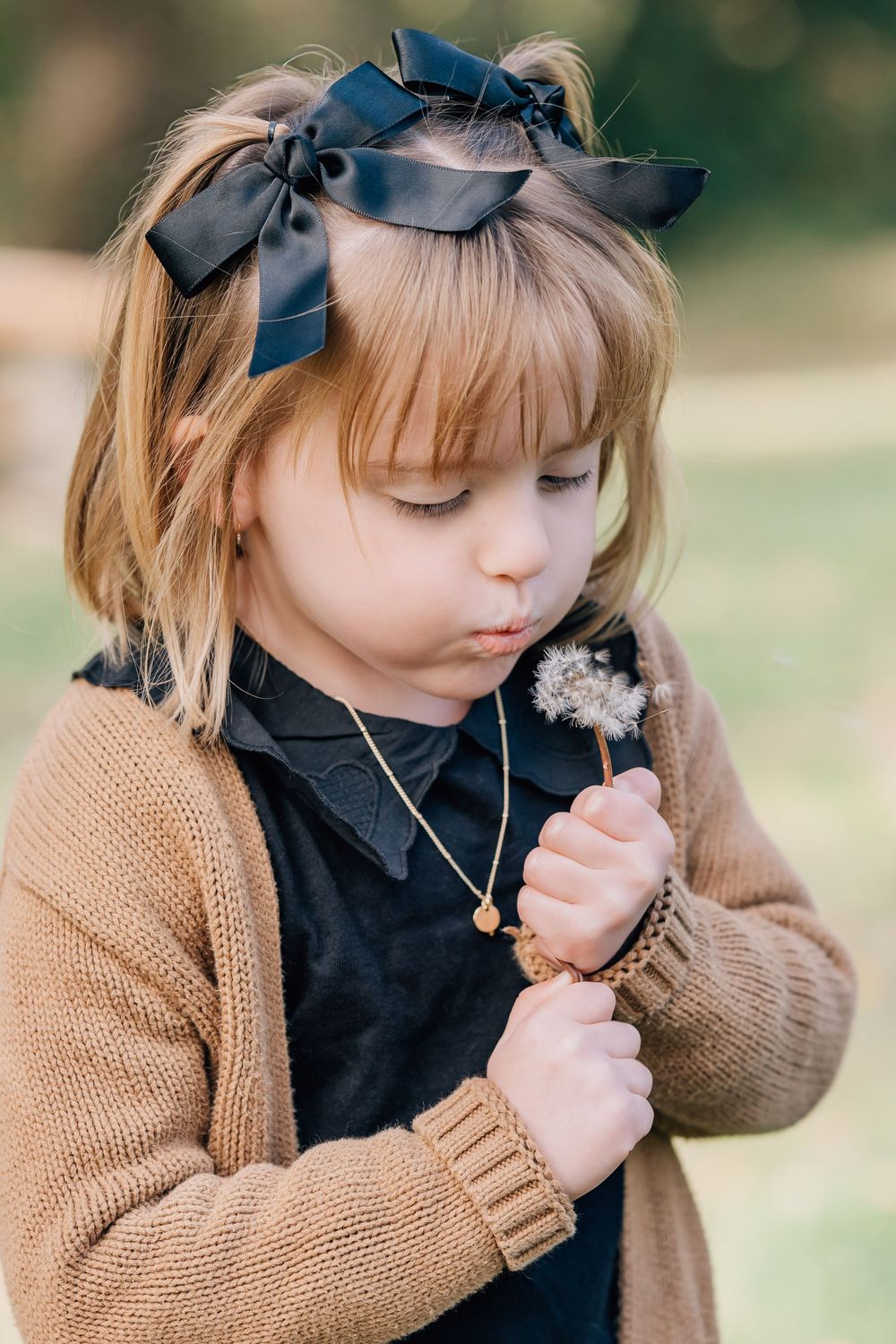 Young girl with black bow blowing dandelion, wearing tan knit cardigan and gold necklace outdoors.