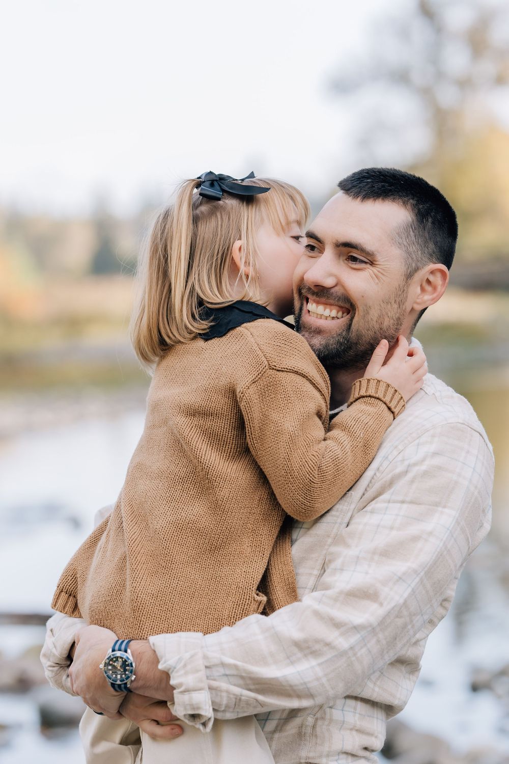 A smiling man holds a young girl in a brown sweater who kisses his cheek outdoors near a pond.